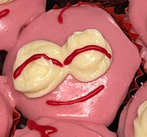 a plate of 7 pink cupcakes, each one uniquely decorated with icing to look like pim. from left to right/top to bottom, the expressions are smiling looking up, smiling and winking, smiling with tongue out, smiling with eyes closed, smiling with mouth open, perplexed, and a neutral front facing pim.